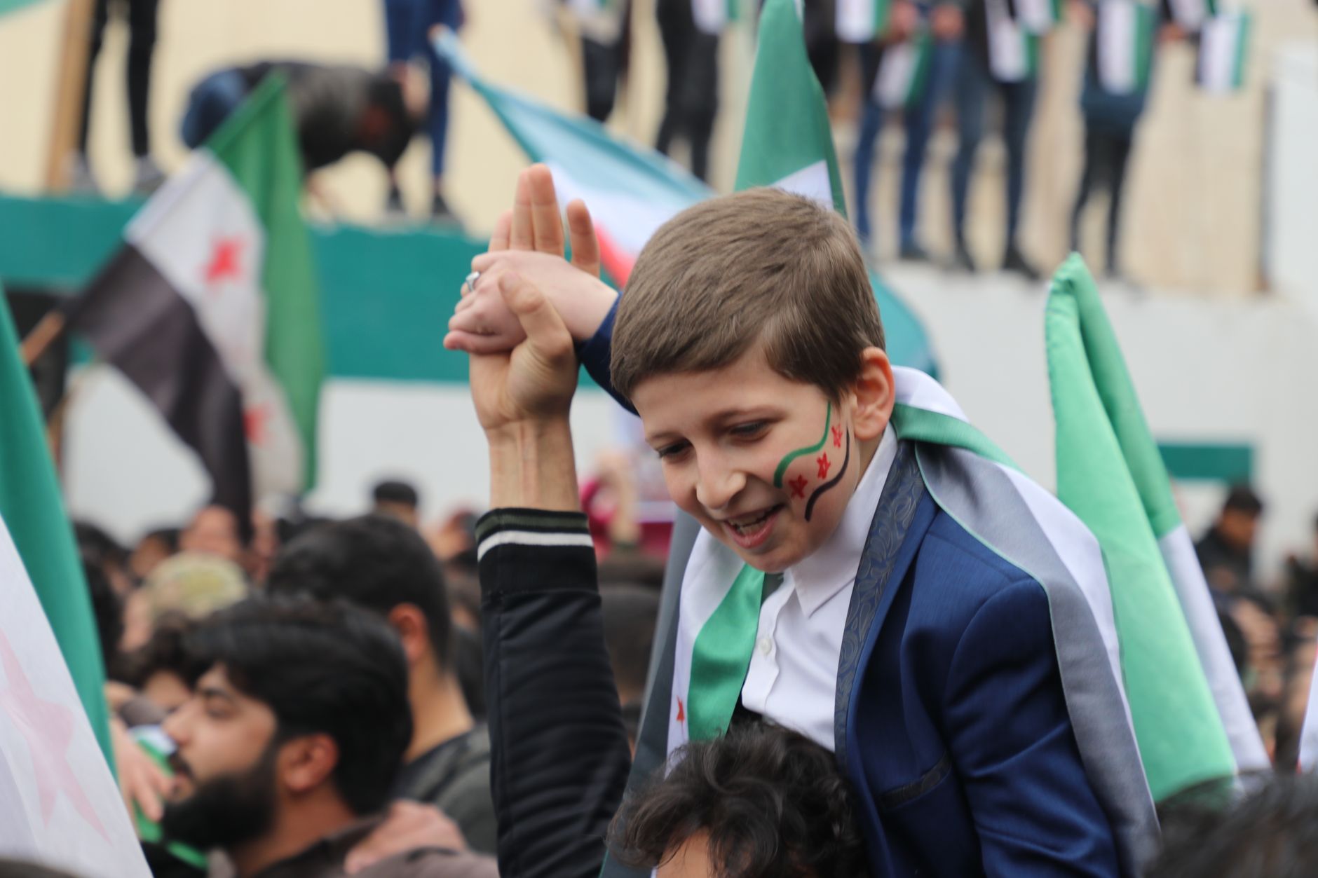 Young Syrian boy with face paint at a vibrant rally, showcasing national pride.
