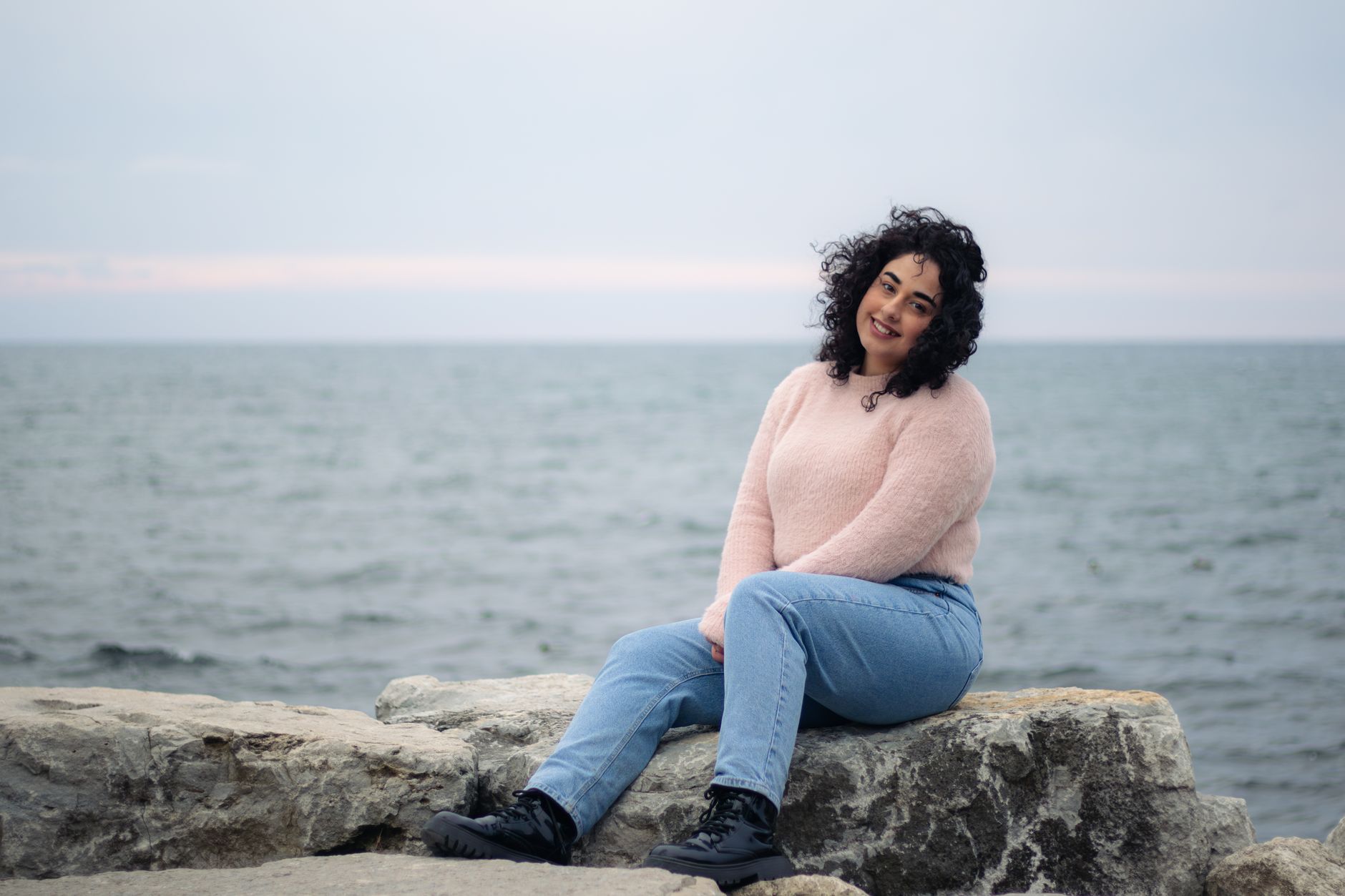 A woman with curly hair sits on rocks by the sea in Toronto, smiling warmly.