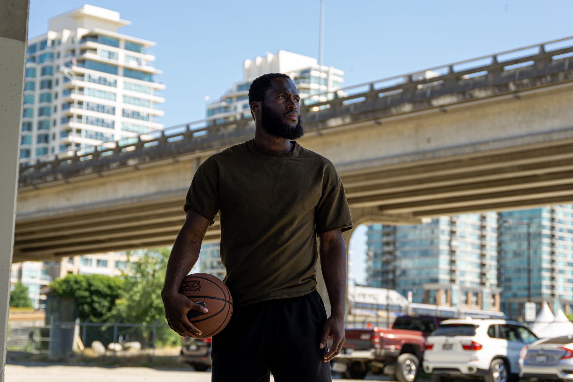 A man holding a basketball stands beneath a city bridge, surrounded by urban architecture and cars.